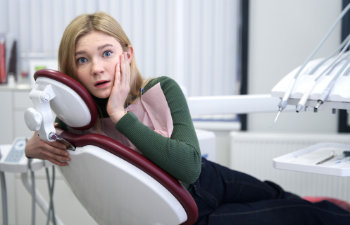 Woman in dental chair looks anxious, touching her cheek, in a dentist's office.