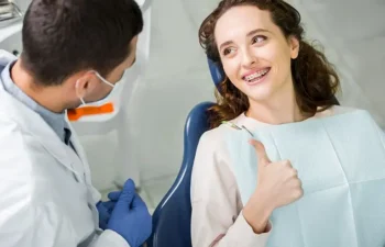 A woman with braces gives a thumbs up while sitting in a dental chair, as a dentist in a white coat and gloves speaks to her.